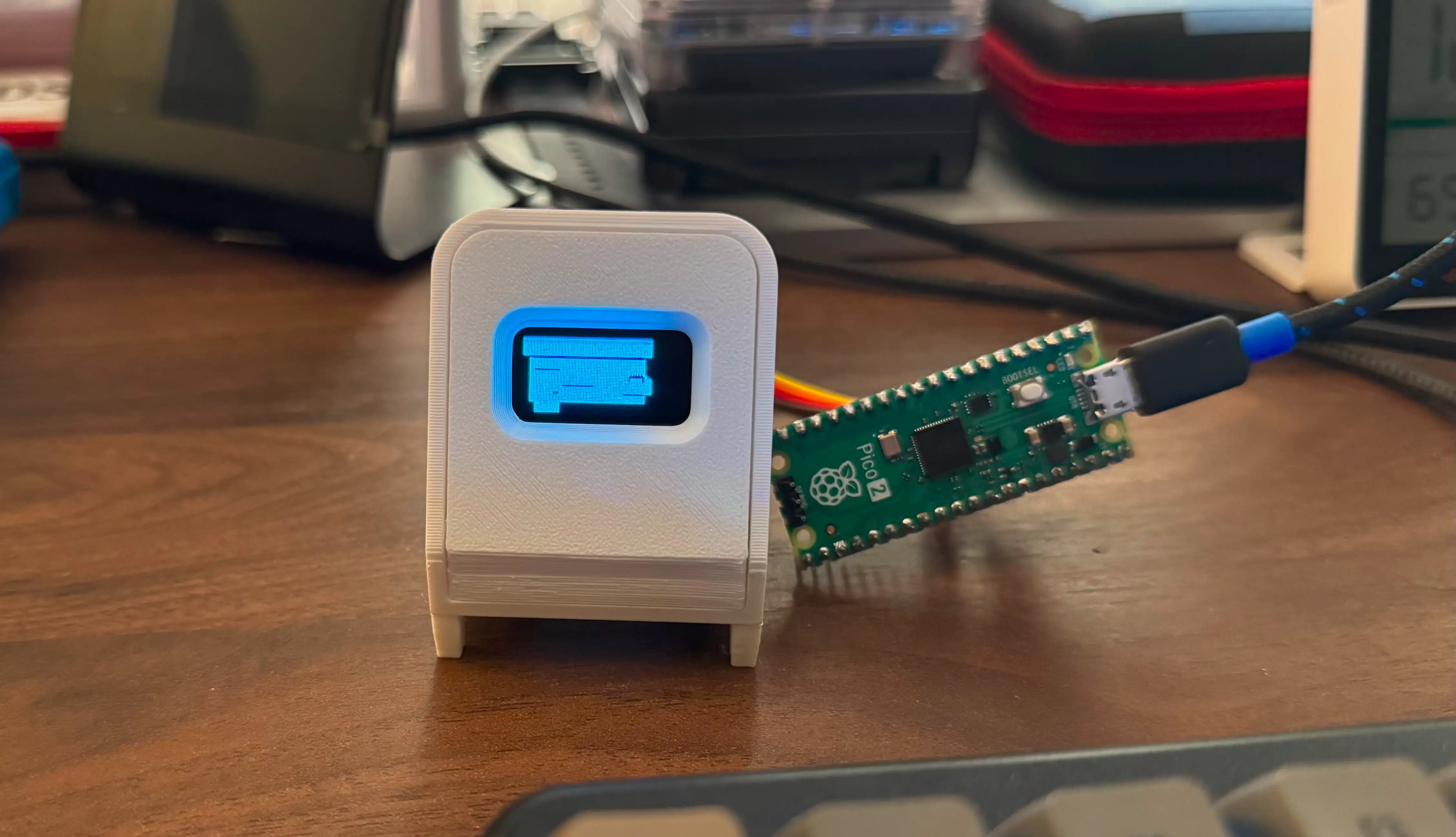 Picture of 3D printed case for a SSD1306 OLED with the screen in it showing the Poststation logo in blue. Beside it is a Pico 2 